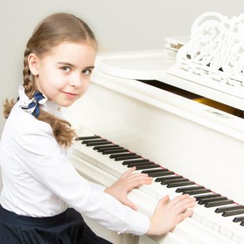 Charming little blonde with long wattled hair in plaits, playing on a white grand piano. In musical school.