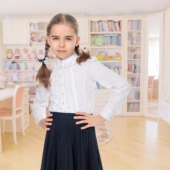 A beautiful little schoolgirl girl in a white blouse and black long skirt, with neatly braided pigtails on her head.She is posing in front of the camera, her hands on her hips.In the children's room.