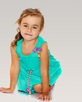 Adorable little blonde girl, dressed in a blue dress standing on her knees. She looks directly into the camera.On a gray background.