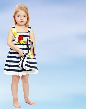 The little blonde girl in a summer striped dress. A girl holds a beach bag.On the background of summer blue sky and fluffy clouds.