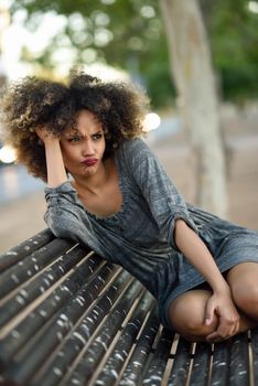 Funny black woman with afro hairstyle sitting on a bench in urban background. Mixed girl wearing casual clothes