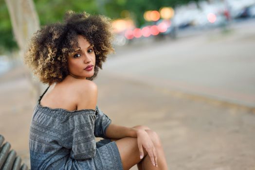 Young black girl with afro hairstyle sitting on a bench in urban background. Mixed woman wearing casual clothes