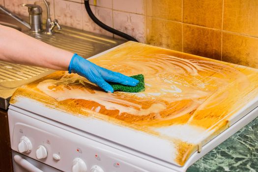 Woman's housewife hand in a household glove washes a dirty coating on a gas stove on kitchen background, close up.