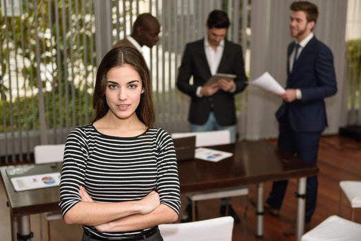 Young businesswoman leader looking at camera with arms crossed in working environment. Group of multiracial people in the background