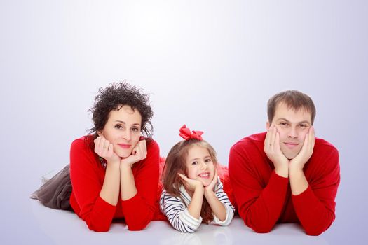 Happy young family dad mom and a little girl in bright red outfits . Family lying on the floor leaning on his hands.Purple gradient background.