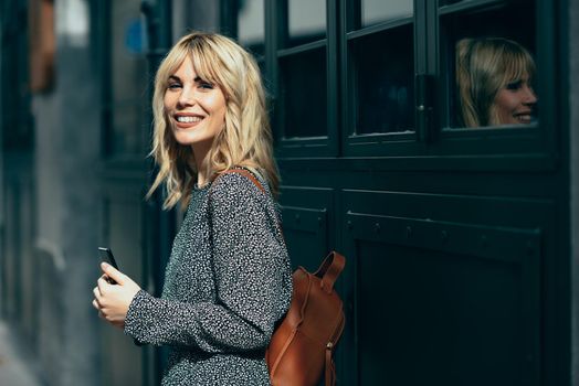 Attractive young caucasian woman looking at her smart phone in urban background. Blonde girl wearing casual clothes outdoors