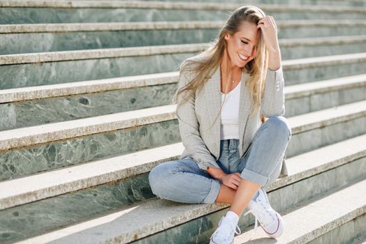 Beautiful young caucasian woman smiling in urban background. Blond girl wearing casual clothes in the street. Female with elegant jacket and blue jeans sitting on stairs.