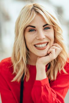 Happy young blond woman sitting on urban background. Smiling blonde girl with red shirt enjoying life outdoors.