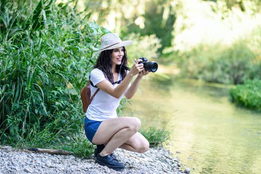 Beautiful hiker young woman taking photographs with a mirrorless camera, wearing straw hat, hiking in the countryside.