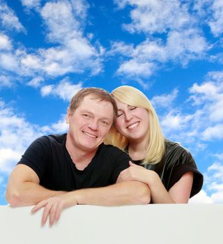 A man and a woman tenderly hugging each other. Peeping over white banner.On the background of summer blue sky and fluffy clouds.
