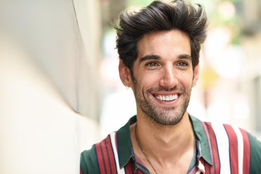 Attractive young man with dark hair and modern hairstyle smiling wearing casual clothes in urban background.