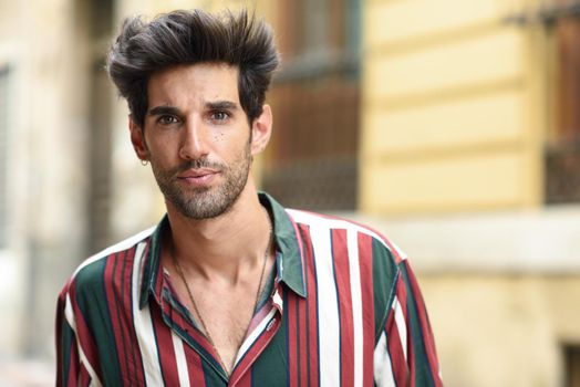 Handsome young man with dark hair and modern hairstyle wearing casual clothes in urban background.