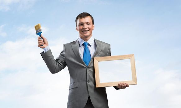 Creative businessman painter holding paint brush and whiteboard in hands. Portrait of happy handsome man in business suit and tie on blue sky background. Ambitions and creativity in business.