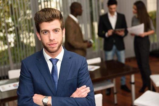 Image of male business leader looking at camera in working environment. Man with arms crossed wearing blue suit and tie.