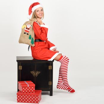 Beautiful happy blonde woman in Santa Claus clothes and striped socks sitting on a locker. Young funny girl with blue eyes carrying a sack of santa claus, isolated on white