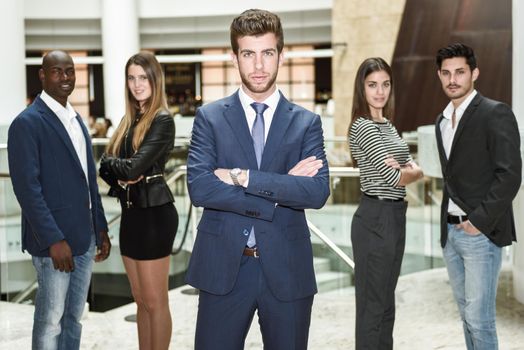 Businessman leader looking at camera with arms crossed in office building. Group of muti-ethnic people in the background