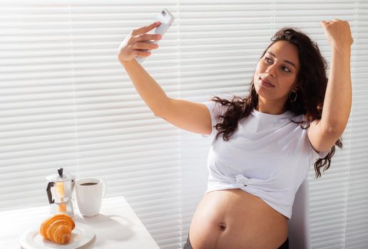 Happy pregnant young beautiful woman talking to mom using video call during morning breakfast. Communication and positive attitude during pregnancy