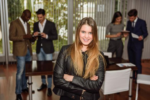 Blonde businesswoman leader looking at camera with arms crossed in working environment. Group of people in the background