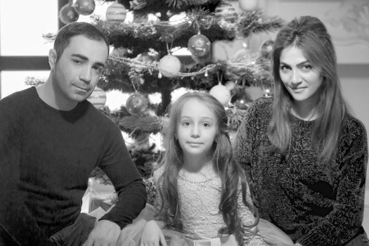 Portrait of loving parents hugging their charming daughter, mom and dad, sitting on a sofa near the Christmas Tree. Against the background of a large panoramic window in the studio. Holidays concept.