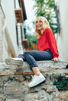 Happy young blond woman sitting on urban background. Smiling blonde girl with red shirt enjoying life outdoors.