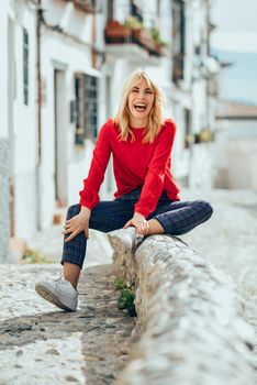 Happy young blond woman sitting on urban background. Laughing blonde girl with red shirt enjoying life outdoors.