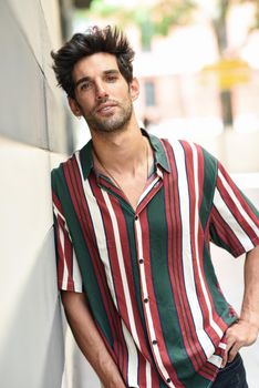 Attractive young man with dark hair and modern hairstyle wearing casual clothes in urban background.