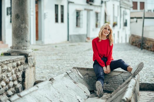 Happy young blond woman sitting on urban background. Smiling blonde girl with red shirt enjoying life outdoors.