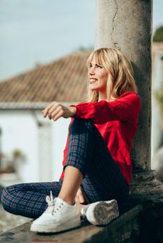 Happy young blond woman sitting on urban background sunbathing in autumn. Smiling blonde girl with red shirt enjoying life outdoors.