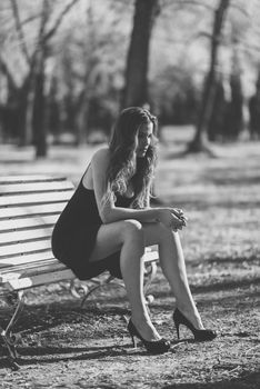 Portrait of a pretty woman sitting in a bench in a urban park wearing black dress and high heels