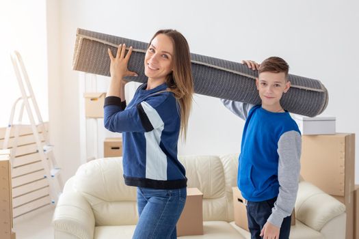 Smiling caucasian mother and a charming son are holding a folded carpet on their shoulders in a new living room intending to spread it in a new apartment. Housewarming concept