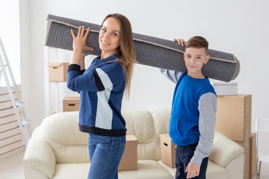 Smiling caucasian mother and a charming son are holding a folded carpet on their shoulders in a new living room intending to spread it in a new apartment. Housewarming concept