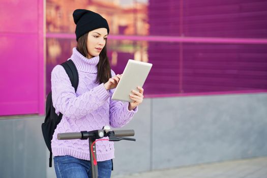 Woman in her twenties with electric scooter using digital tablet outdoors. Lifestyle concept.