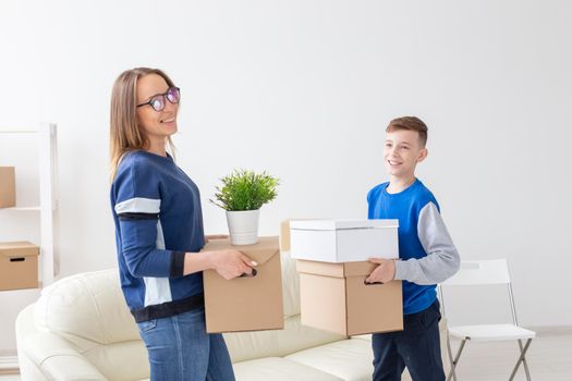 Young single mother and a playful little son are holding a box with things and a flower in a pot standing in the living room in a new apartment. Housewarming and new housing concept