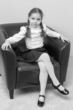 A sweet blonde with long wattled hair in pigtails sits on a brown leather chair and puts her foot on her leg.Black-and-white photo. Retro style.