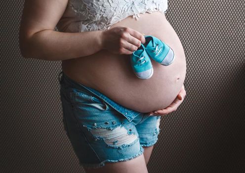 Hands of a pregnant girl hold belly and small baby booties, awaiting the birth of a baby.