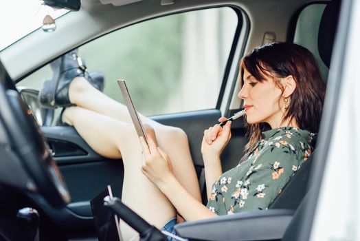 Young woman writing in a notebook with a pen in a white car pulling her feet out the window. Female wearing flowered shirt and shorts.
