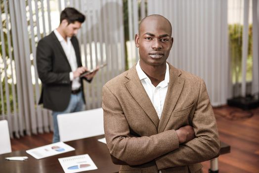 Confident businessman. Cheerful young African man in formalwear keeping arms crossed and smiling while his caucasian colleague standing on background