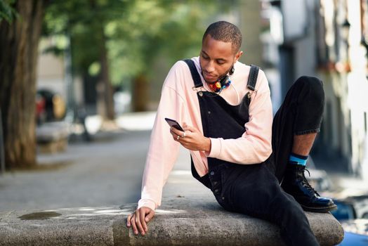 Young black man with headphones sitting in urban street looking at his smart phone. Lifestyle concept.
