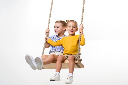 Happy kids swinging on rope swing against white background. Cute smiling brother and sister having fun on summer holidays, playing in playground or kindergarten. Happy childhood concept