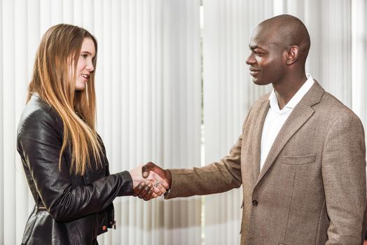 Black businessman shaking hands with a blonde caucasian young woman wearing suit in a office