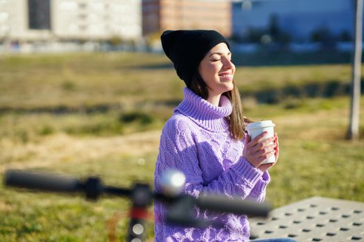 Female student with electric scooter taking a coffee break enjoying the winter sunlight with her eyes closed. Lifestyle concept.