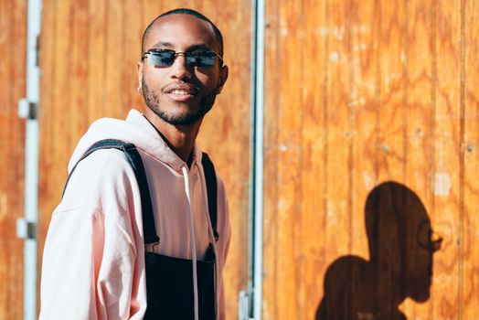 Young black man wearing casual clothes and sunglasses, smiling against a wooden background. Millennial african guy with bib pants outdoors