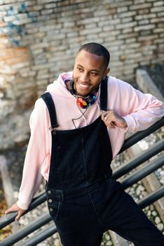 Young black man wearing casual clothes smiling on urban stairs. Happy African guy with bib pants outdoors