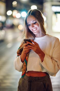 Blonde woman looking at her smartphone in the street. Defocused city lights at the background. Pretty girl with pigtail hairstyle at night.
