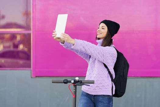 Woman in her twenties with electric scooter taking a selfie with a digital tablet outdoors. Lifestyle concept.