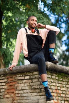 Young black man wearing casual clothes and headphones sitting on urban background. Happy African guy with bib pants outdoors