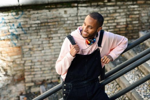 Young black man wearing casual clothes smiling on urban stairs. Happy African guy with bib pants outdoors