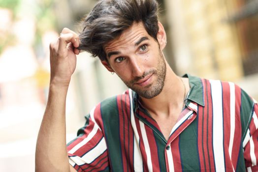 Attractive young man with dark hair and modern hairstyle wearing casual clothes in urban background.