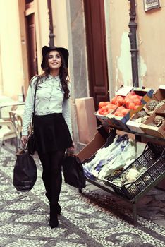 Young woman with shopping bags walking near a fruit's shop. Wearing skirt, shirt and hat.