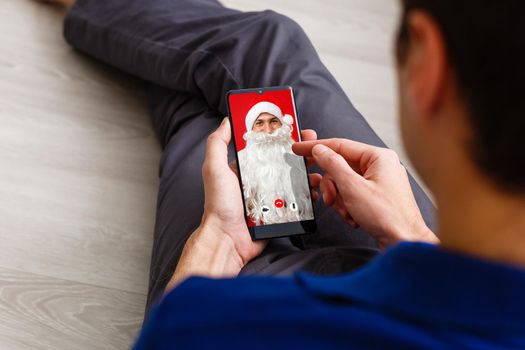 Christmas online congratulations. Surprised, shocked man wearing medical mask talking on the phone with friends and parents. christmas tree bokeh background